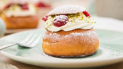 A close-up of a freshly baked bun topped with whipped cream, raspberries, and pistachios, dusted with powdered sugar on a green plate.