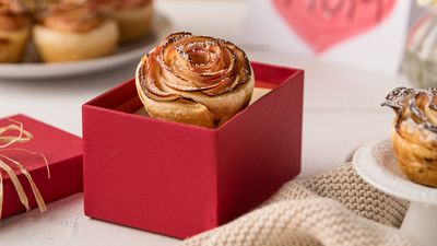 A beautifully crafted rose-shaped pastry in a red gift box, surrounded by a cozy setting with a knitted cloth and a heart-shaped card.