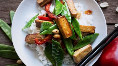 A plate of stir-fried tofu and vegetables, including snap peas and red bell pepper, served over white rice with peanuts.