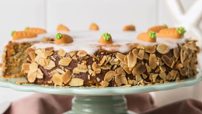 A festive carrot cake topped with almond slices, white icing, and small fondant carrot decorations on a pale green cake stand.