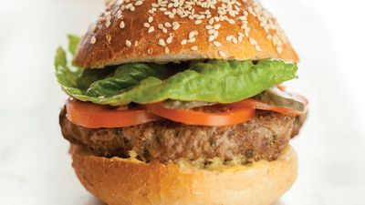 Close-up of a sesame seed bun burger, featuring a beef patty, lettuce, tomato, and pickles. Brightly colored and appetizing.