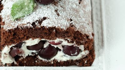 A rich chocolate roll cake dusted with powdered sugar, filled with creamy frosting and juicy cherries, garnished with a mint leaf.