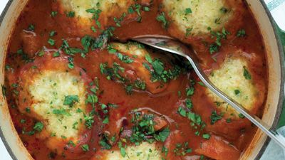 A close-up of fluffy dumplings in a rich, savory tomato broth, garnished with fresh parsley. A spoon rests in the dish.