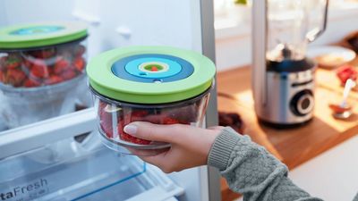 A person placing a glass container with a green lid filled with strawberries into a refrigerator.