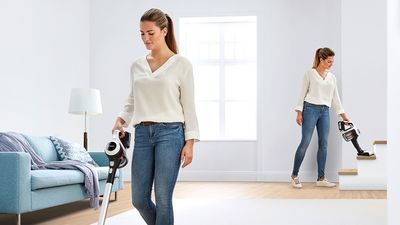 A woman in a white blouse and jeans uses a handheld vacuum to clean a bright, modern living space with wooden flooring and a cozy couch.