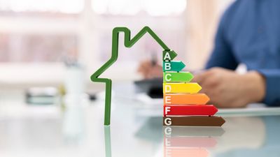 A colorful energy efficiency rating scale beside a green house outline, with a person sitting at a desk in the background.