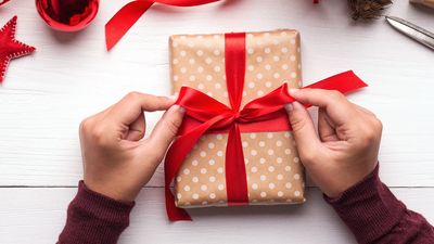 Two hands tying a red ribbon around a polka-dotted gift box on a white wooden surface, surrounded by festive decorations.