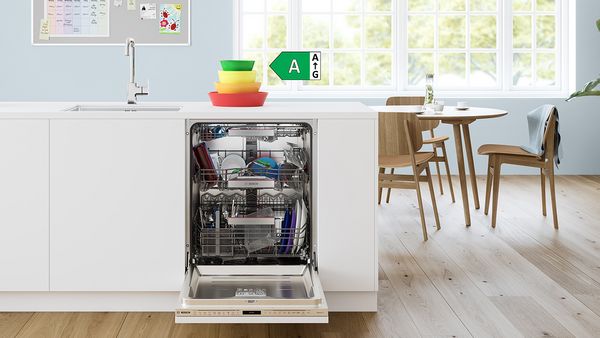 Open dishwasher in a kitchen full of colourful crockery and utensils, next to a dining table with wooden chairs and sunlight.