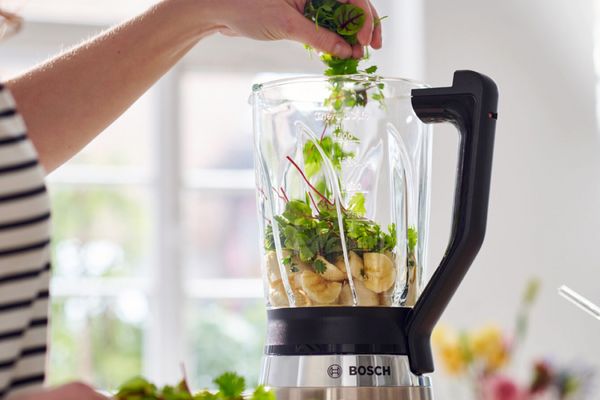 A woman puts vegetables into a BOSCH blender.