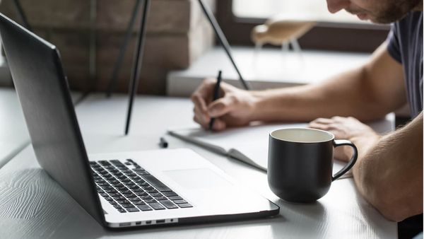 Person writing in a notebook next to a laptop and a cup of coffee on a desk.