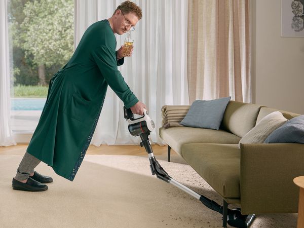 Man in groene jas stofzuigen woonkamer met groot raam en bank.