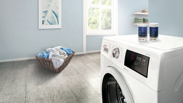 A modern washing machine in a bright laundry room, alongside a basket of laundry and laundry products on a countertop.