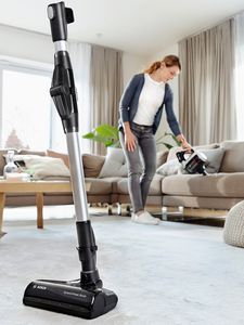 A woman is vacuuming a light-coloured carpet near a couch with a cordless vacuum cleaner, while another vacuum cleaner is nearby. 