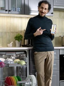 A person in a kitchen is standing next to a dishwasher full of colourful dishes, pointing to a smartphone in her hand. 