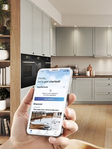 
       Людина ніжно тримає телефон, зосереджена на екрані Home Connect, на фоні затишна кухня.
      