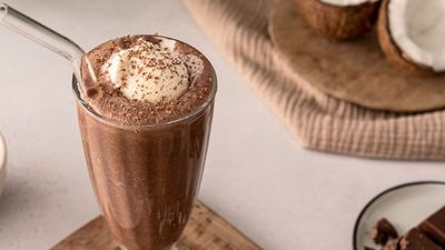 A frosty chocolate milkshake topped with whipped cream and chocolate shavings, served with coconut halves in a cozy setting.