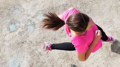 Aerial view of a woman jogging outdoors in a bright pink shirt and black leggings, with long brown hair flowing behind her.