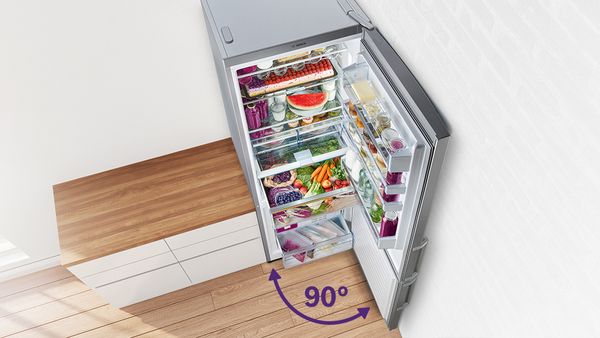 A modern refrigerator filled with fresh produce and drinks, open at a 90-degree angle, beside a wooden countertop 