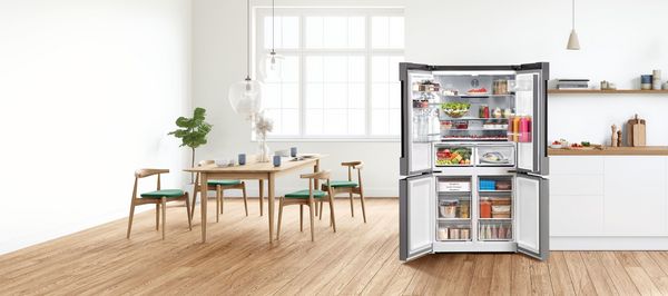 A hand holds a smartphone showing the contents of a fridge, which is visible in the bright kitchen in the background.