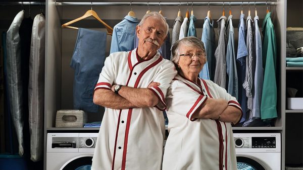 Two fit retirees in matching white tops stand back to back in an elegant laundry room.
