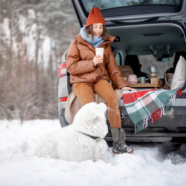 Person sitting on a car trunk in the snow, enjoying a drink with a dog nearby. 