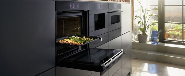 Moderne keuken met oven, magnetron en groenten op bakplaat.