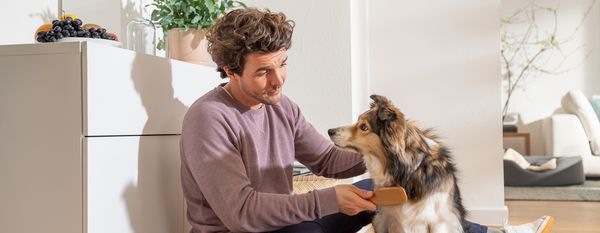 Man aaiende hond in huis, glimlachend, omgeven door planten en meubels.