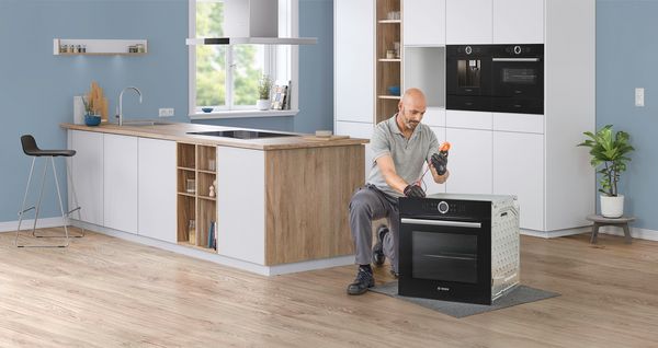 Modern living room with a gray sofa, coffee cup on a wooden table, and a view into a bright kitchen featuring a refrigerator and large windows.