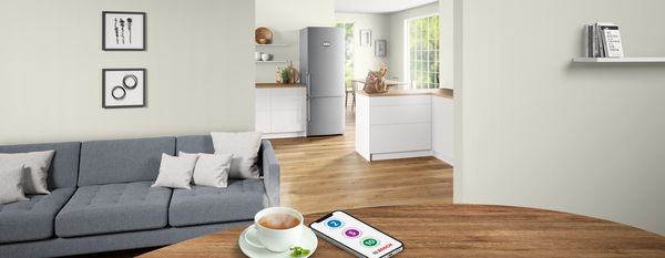 Modern living room with a gray sofa, coffee cup on a wooden table, and a view into a bright kitchen featuring a refrigerator and large windows.
