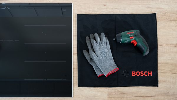 A Bosch induction hob with gloves and a drill placed on a black cloth beside it. 
