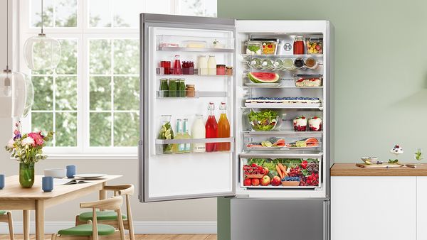 In a bright, modern kitchen, the open door of a fridge freezer reveals an interior full of colourful fresh produce, including bottles of syrups, smoothies and preserves in the door.