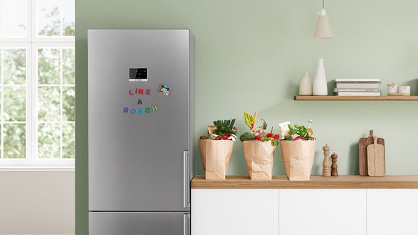 A stainless steel fridge with "Like a Bosch" written on it in coloured fridge magnets stands next to a counter where three bags of fresh produce are waiting to be unpacked.