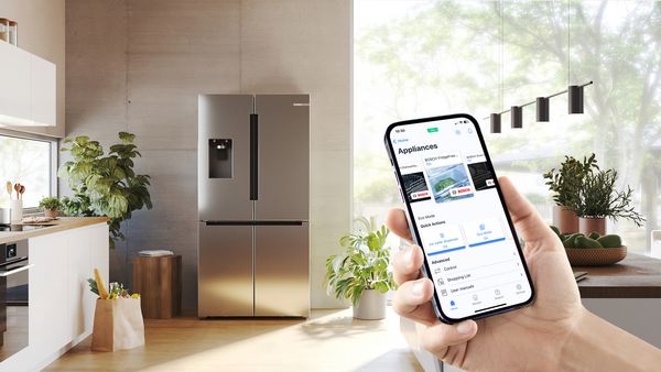 A hand holds a smartphone, which displays the contents of a fridge that can be seen in the warm, bright kitchen area in the background.