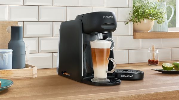 TASSIMO FINESSE in the colour lupine blue brewing a Latte Macchiato on a kitchen worktop and a plate of pancakes next to it.